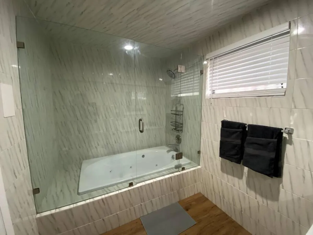 A modern, spa-inspired bathroom with a glass-enclosed shower, inviting jacuzzi tub, sleek modern fixtures, and warm wooden flooring. The space is flooded with natural or gentle artificial light, highlighting the room’s fresh and luxurious atmosphere.