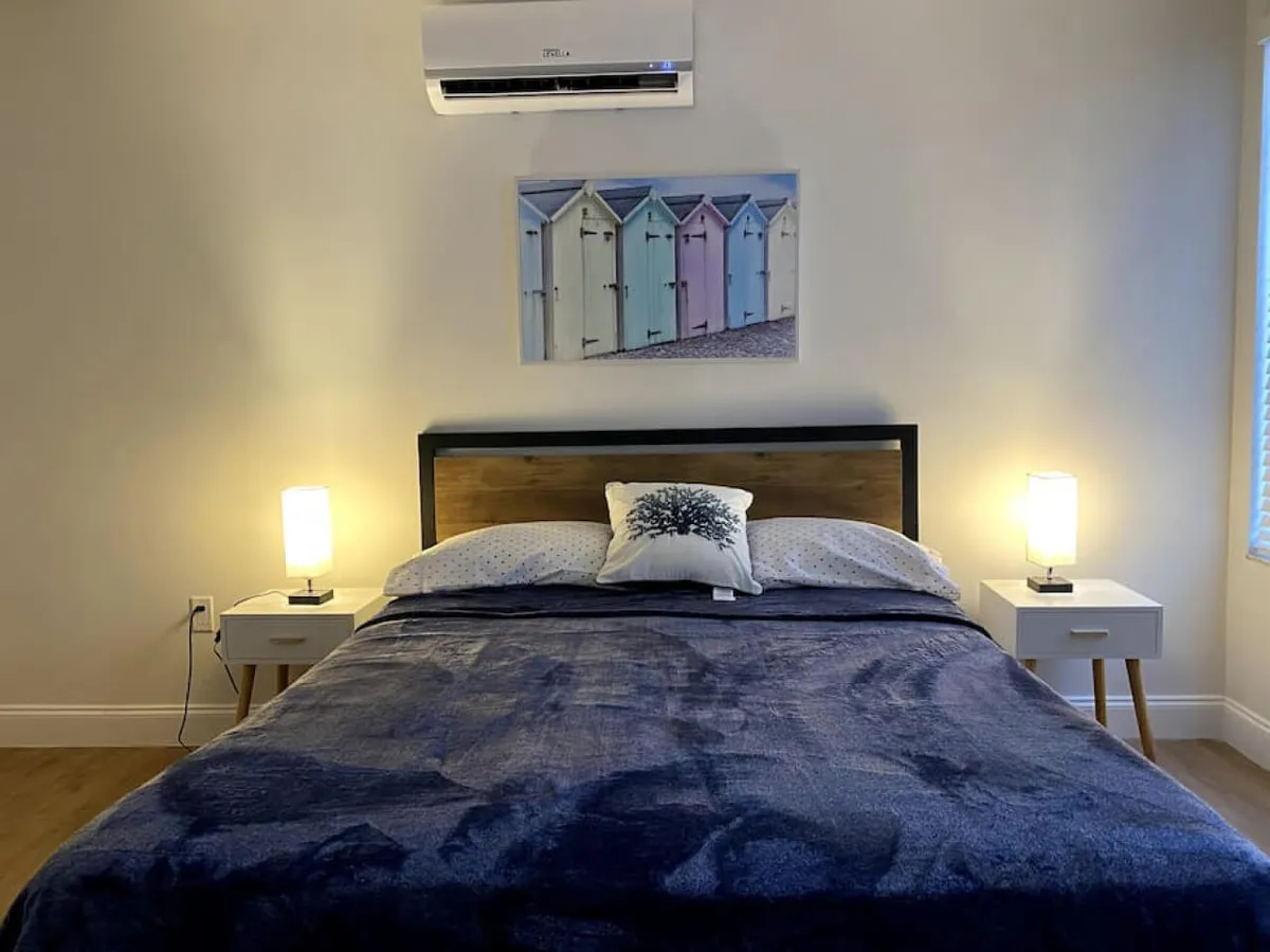 The image showcases a serene bedroom with a plush bed dressed in dark blue bedding, flanked by two modern bedside tables and softly glowing lamps. Above the bed, pastel beach hut wall art adds a coastal Miami vibe, and an air conditioning unit promises cool comfort. The room exudes a peaceful, inviting atmosphere perfect for relaxation.