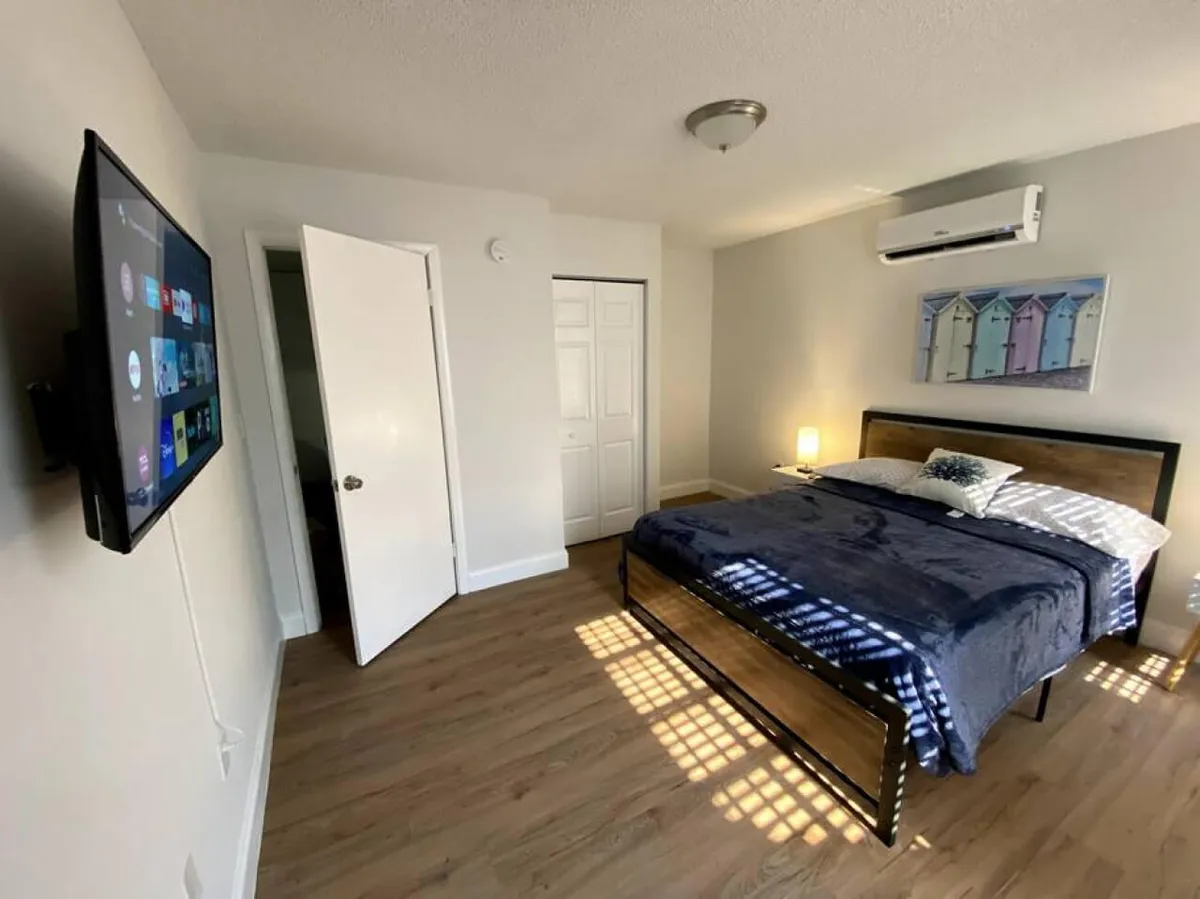 A spacious bedroom featuring a king-size bed with a dark blue blanket and patterned pillows. Natural light filters through window blinds, illuminating the wooden floor. Modern wall art depicting colorful beach huts hangs above the bed, and a wall-mounted flat-screen smart TV is positioned opposite for entertainment. The room feels inviting and restful, ideal for relaxation and comfort.