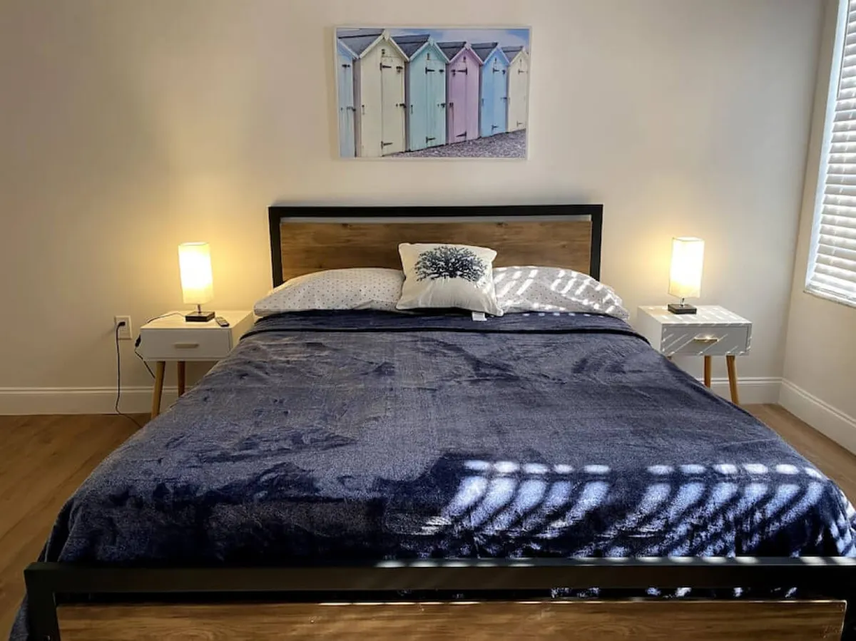 A tranquil bedroom scene with a wooden bed frame topped with dark blue bedding, flanked by two bedside tables with lamps. The wall art above the bed features pastel-colored beach huts, and natural light filters in from a nearby window, creating a soothing ambiance perfect for unwinding.