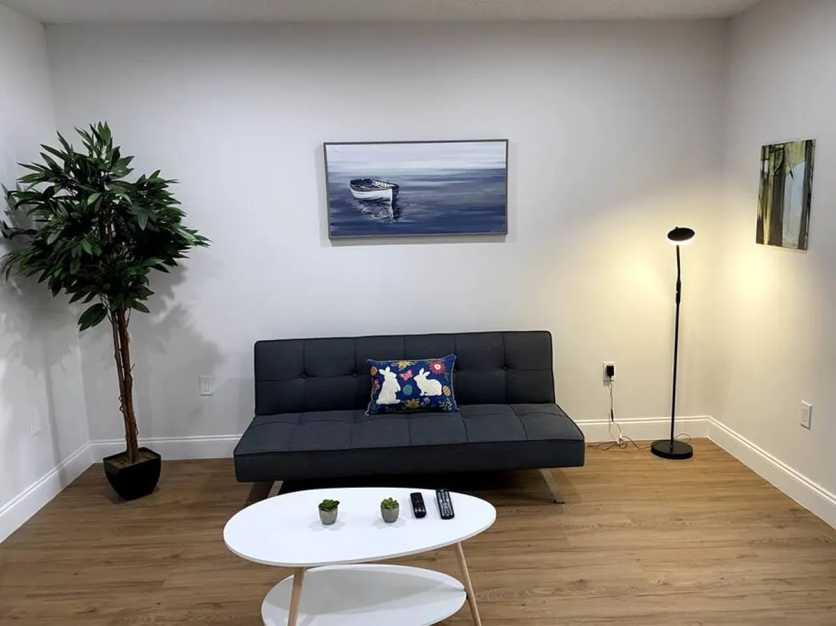 A calm, cozy living room featuring a dark gray sofa accented by a decorative pillow, a white oval coffee table with remotes and small plants, a floor lamp providing warm light, and a large potted plant in the corner. Natural light fills the space, highlighting the comfortable seating area ideal for relaxing, socializing, or enjoying TV after a day in the city.