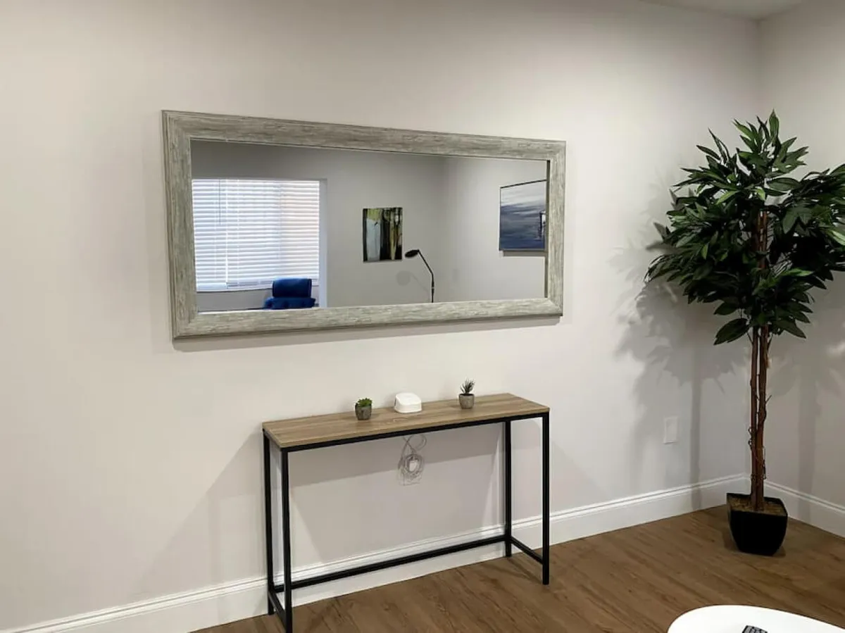 A welcoming entryway with a large framed wall mirror, a console table holding decorative plants and a small device, and a tall potted artificial plant. The scene is neat and uncluttered, creating a sense of order and making it easy to imagine seamless arrivals and departures.