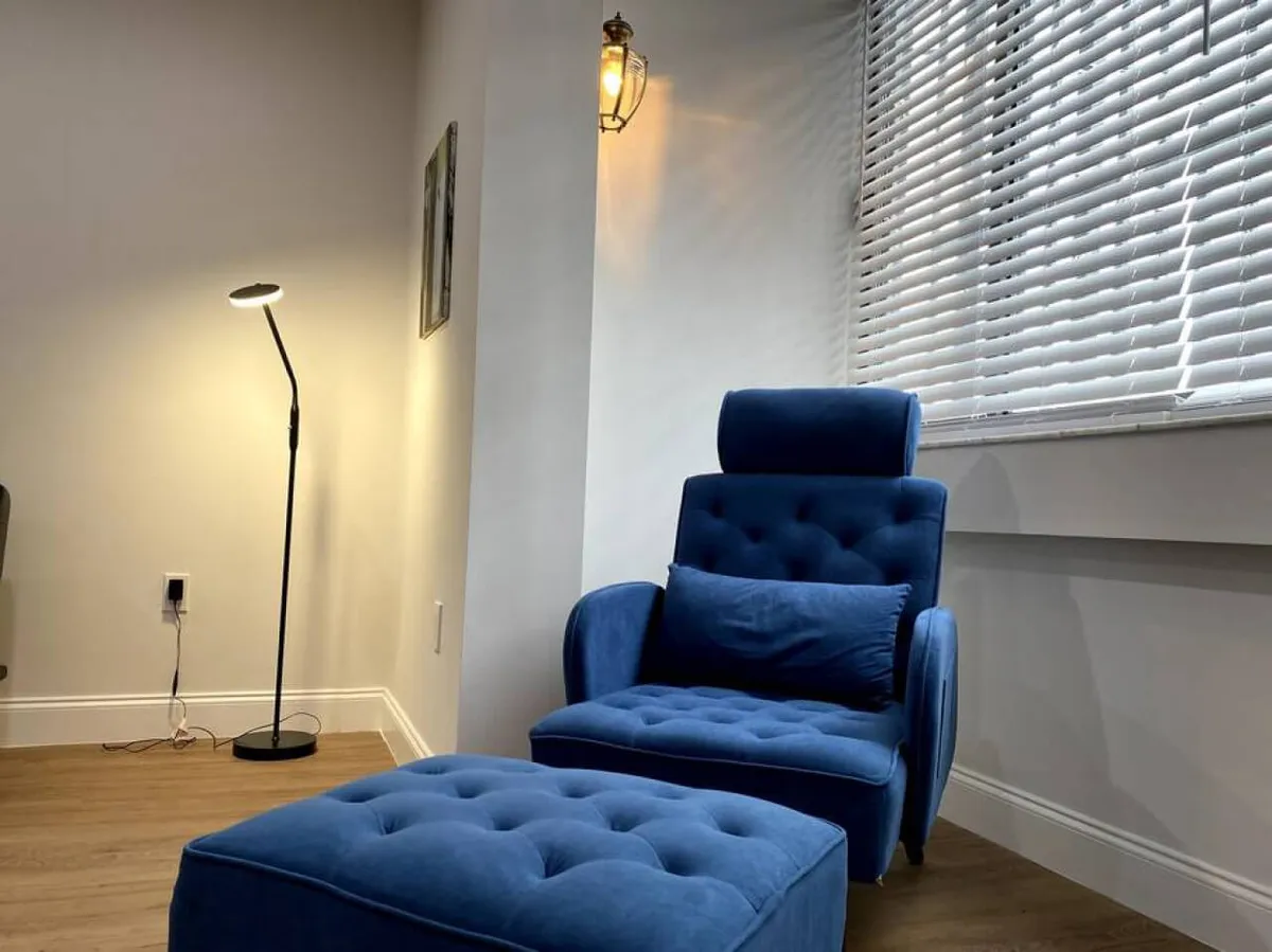 A cozy corner featuring a blue tufted armchair and matching ottoman, bathed in natural light from a nearby window. A floor lamp adds a warm glow, creating a comfortable spot for relaxing or enjoying a quiet moment with coffee.