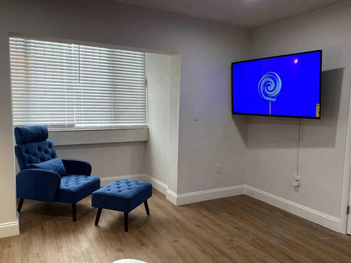 A modern living space with a blue upholstered armchair and matching ottoman, a large wall-mounted smart TV, and a window with white blinds letting in abundant natural light. The setup invites guests to unwind, read, or enjoy streaming entertainment, emphasizing both comfort and modern amenities.