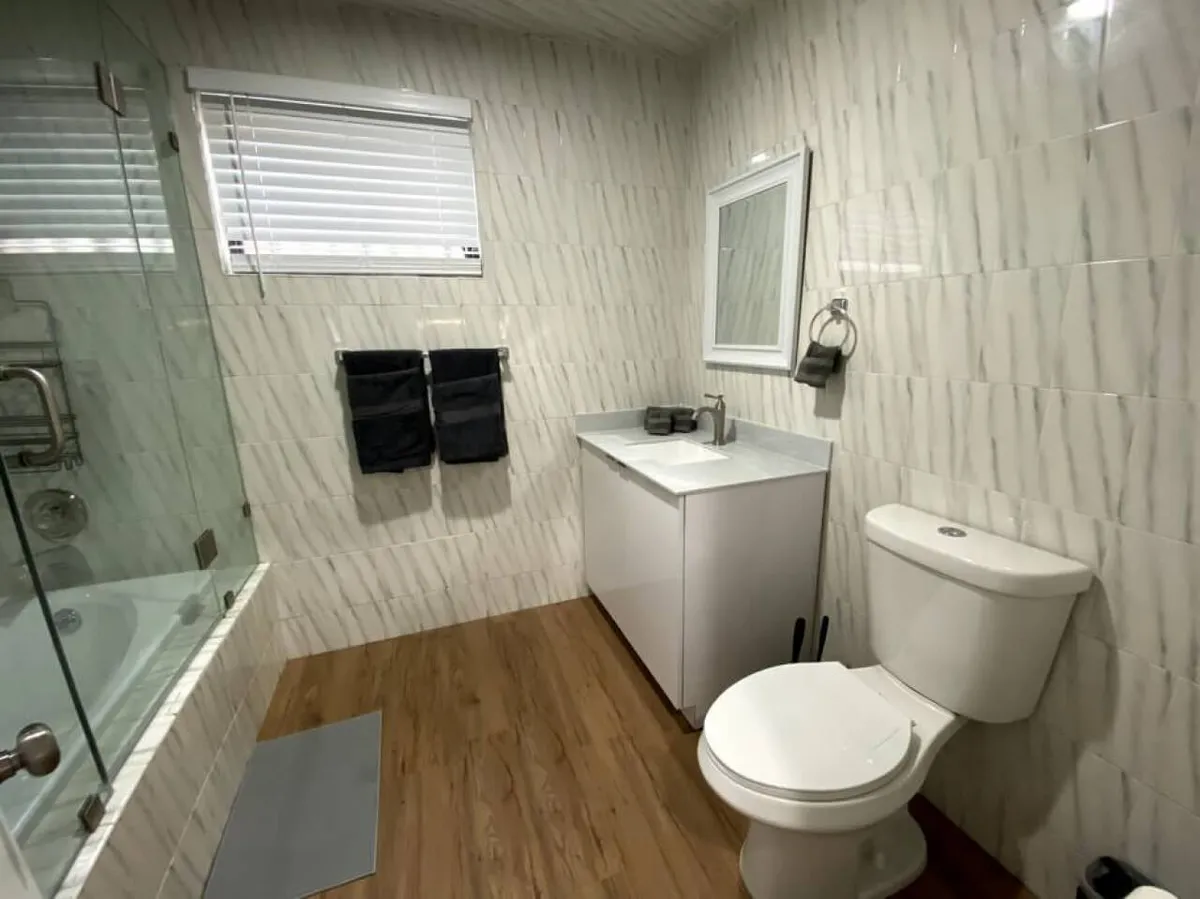 This image showcases a modern bathroom featuring a bathtub with a glass partition, a white vanity with an integrated sink and mirror, a toilet, and a window with blinds. The space is clean, bright, and thoughtfully designed, reflecting comfort and practicality.