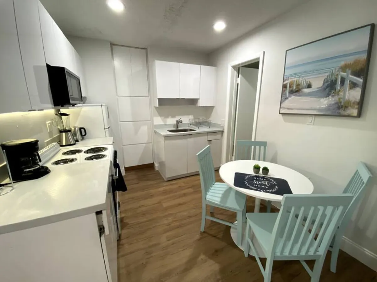 This image showcases a bright, inviting kitchen with white cabinetry and countertops. A small round dining table with pastel blue chairs sits nearby, and kitchen appliances—including a coffee maker and blender—are visible. Beach-themed wall art enhances the Miami ambiance.